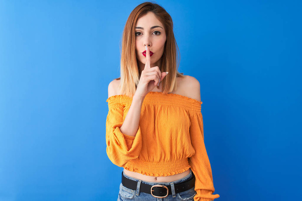 Belle femme rousse portant un t-shirt orange décontracté debout sur un fond bleu isolé demandant à être tranquille avec le doigt sur les lèvres. Silence et concept secret
. - Photo, image