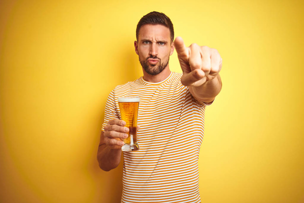 junger gutaussehender Mann trinkt ein Pint Bier auf isoliertem gelben Hintergrund und zeigt mit dem Finger auf die Kamera und auf dich, Handzeichen, positive und selbstbewusste Geste von vorne - Foto, Bild