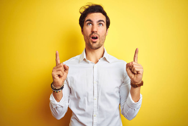 Young handsome businessman wearing elegant shirt standing over isolated yellow background amazed and surprised looking up and pointing with fingers and raised arms. - Foto, imagen