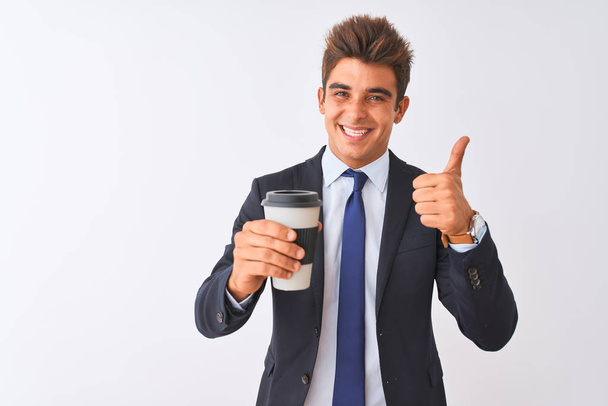 Jonge knappe zakenman in pak met koffie over geïsoleerde witte achtergrond blij met grote glimlach doet ok teken, duim omhoog met vingers, uitstekend teken - Foto, afbeelding