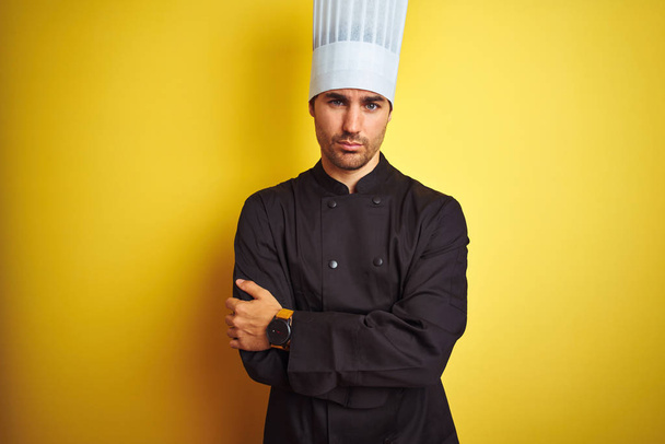 Młody szef kuchni w mundurze i kapeluszu stojący nad odizolowanym żółtym tle sceptyczny i nerwowy, dezaprobata wyraz twarzy z skrzyżowanymi ramionami. Osoba negatywna. - Zdjęcie, obraz