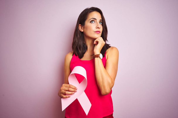 Jovem bela mulher segurando fita de câncer sobre rosa isolado fundo sério rosto pensando em questão, ideia muito confusa
 - Foto, Imagem