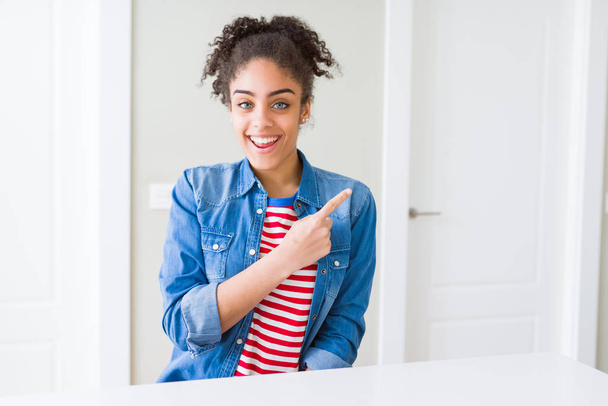 Beautiful young african american woman with afro hair wearing casual denim jacket cheerful with a smile of face pointing with hand and finger up to the side with happy and natural expression on face - Photo, Image