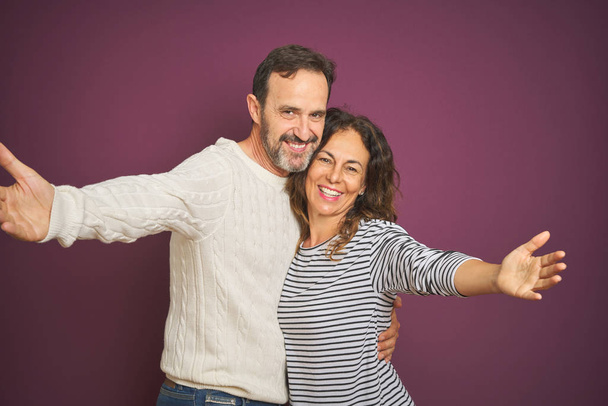 Beau couple d'âge moyen portant un pull d'hiver sur fond violet isolé regardant la caméra souriant à bras ouverts pour un câlin. Expression joyeuse embrassant le bonheur
. - Photo, image