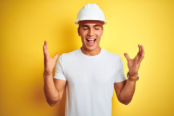Mladý pohledný muž ve stavební helmě na žlutém osamělém pozadí oslavující šílený a šílený úspěch se zdviženýma rukama a zavřenýma očima křičící vzrušeně. Koncept vítěze - Fotografie, Obrázek