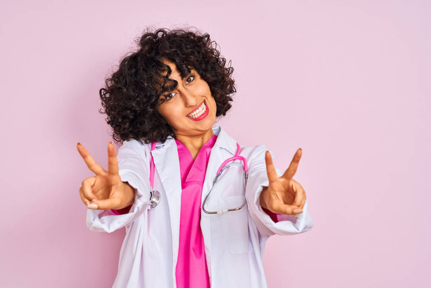 junge arabische Ärztin mit lockigem Haar, Stethoskop über isoliertem rosa Hintergrund, lächelnd in die Kamera blickend, die Finger beim Siegeszeichen zeigend. Nummer zwei. - Foto, Bild