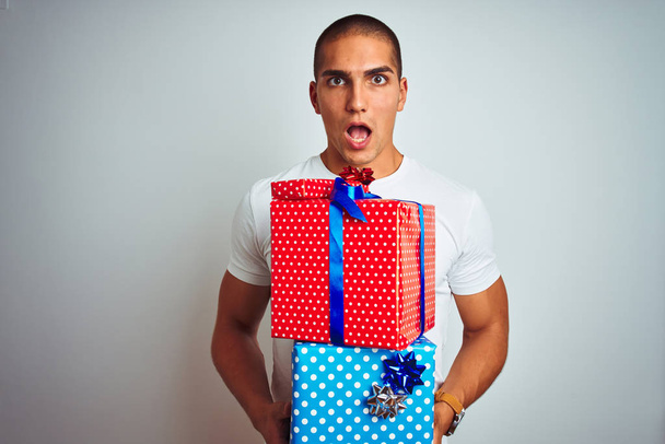 Joven hombre guapo sosteniendo regalos de cumpleaños sobre fondo blanco aislado asustado en shock con una cara sorpresa, asustado y excitado con expresión de miedo
 - Foto, Imagen