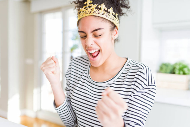 頭の上に黄金の女王の冠を身に着けている若いアフリカ系アメリカ人の少女は非常に満足し、腕を上げて勝者のジェスチャーを行う興奮し、笑顔と成功のために叫んでいます。お祝いのコンセプト. - 写真・画像