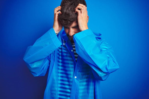 junger gutaussehender Mann im Regenmantel, der vor isoliertem blauen Hintergrund steht und unter Kopfschmerzen leidet, verzweifelt und gestresst wegen Schmerzen und Migräne. Hände auf den Kopf. - Foto, Bild