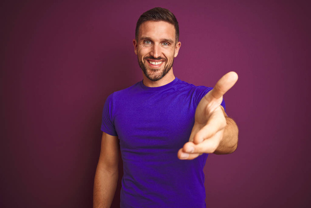 Giovane uomo che indossa casual viola t-shirt su sfondo lilla isolato sorridente amichevole offrendo stretta di mano come saluto e accogliente. Attività di successo
. - Foto, immagini