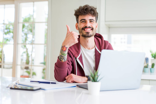 Young student man using computer laptop and notebook happy with big smile doing ok sign, thumb up with fingers, excellent sign - Φωτογραφία, εικόνα