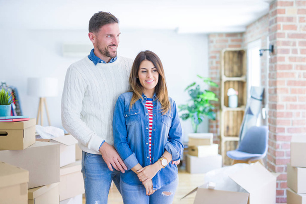 Young beautiful couple standing at new home around cardboard boxes looking away to side with smile on face, natural expression. Laughing confident. - Fotografie, Obrázek