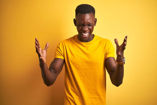 Nuori afrikkalainen amerikkalainen mies päällään rento t-paita seisoo yli eristetty keltainen tausta juhlii hullu ja hullu menestystä kädet kohotetaan ja suljetut silmät huutaa innoissaan. Voittajan käsite
 - Valokuva, kuva