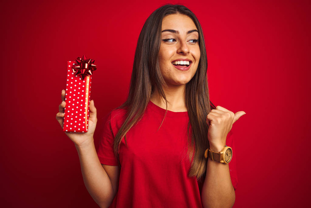 Νεαρή όμορφη γυναίκα κρατώντας δώρο γενεθλίων στέκεται πάνω από απομονωμένο κόκκινο φόντο που δείχνει και δείχνει με τον αντίχειρα μέχρι το πλάι με χαρούμενο πρόσωπο χαμογελώντας - Φωτογραφία, εικόνα