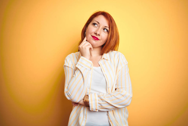 Beautiful redhead woman wearing striped shirt standing over isolated yellow background with hand on chin thinking about question, pensive expression. Smiling with thoughtful face. Doubt concept. - Фото, зображення