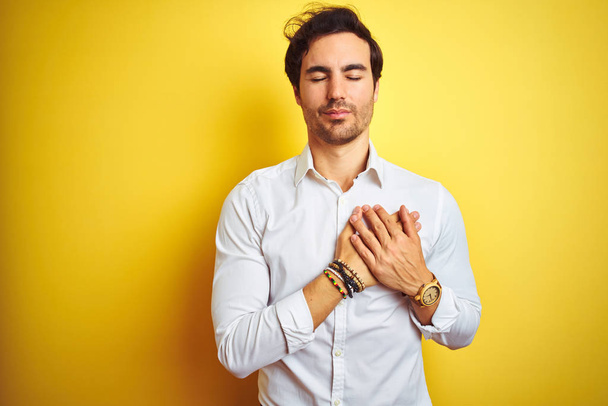 Νέος όμορφος επιχειρηματίας φορώντας κομψό πουκάμισο στέκεται πάνω από απομονωμένο κίτρινο φόντο χαμογελώντας με τα χέρια στο στήθος με κλειστά μάτια και ευγνώμων χειρονομία στο πρόσωπο. Έννοια υγείας. - Φωτογραφία, εικόνα