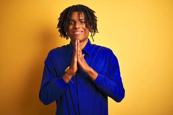Afro worker man with dreadlocks wearing mechanic uniform over isolated yellow background praying with hands together asking for forgiveness smiling confident. - Photo, Image