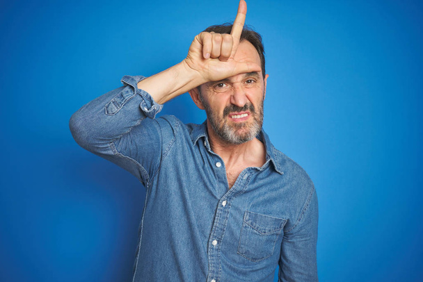 Handsome middle age senior man with grey hair over isolated blue background making fun of people with fingers on forehead doing loser gesture mocking and insulting. - Photo, Image