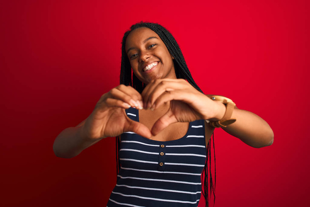 Giovane donna afroamericana che indossa t-shirt a righe in piedi sopra isolato sfondo rosso sorridente in amore che mostra il simbolo del cuore e la forma con le mani. Concetto romantico
. - Foto, immagini