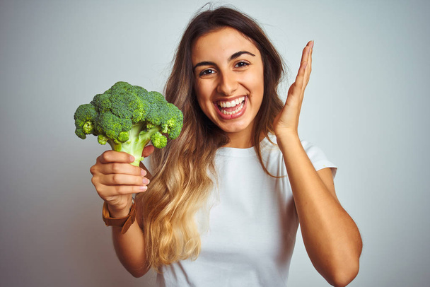 Young beautiful woman eating broccoli over grey isolated background very happy and excited, winner expression celebrating victory screaming with big smile and raised hands - 写真・画像