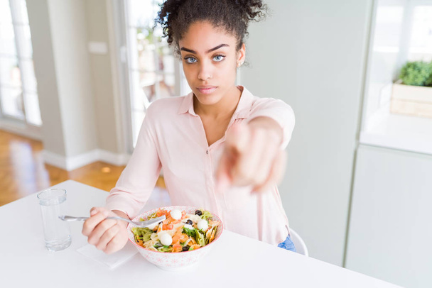 Jonge african american vrouw eten gezond pastasalade met de vinger te wijzen naar de camera en u, hand teken, positieve en zelfverzekerd gebaar van de voorkant - Foto, afbeelding