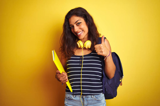 Student kobieta nosi bakcpack notebooka słuchawki na odizolowanym żółtym tle szczęśliwy z dużym uśmiechem robi znak ok, kciuk do góry palcami, doskonały znak - Zdjęcie, obraz
