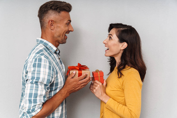 Cute optimistic adult loving couple isolated over grey wall background holding gifts for each other. - Foto, imagen