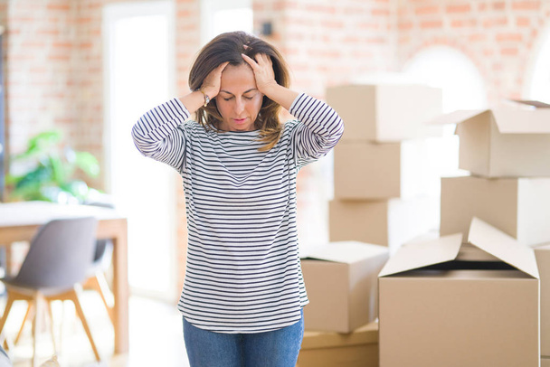 Middelbare leeftijd vrouw verhuizen naar een nieuw huis arround kartonnen dozen die lijden aan hoofdpijn wanhopig en gestrest omdat pijn en migraine. Handen op het hoofd. - Foto, afbeelding