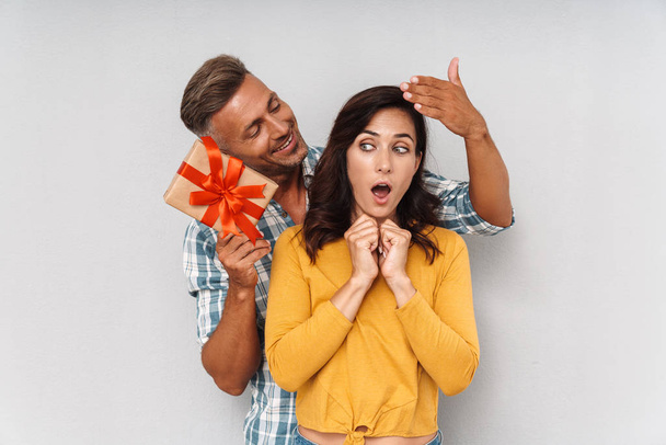 Smiling cheerful man give a present to his shocked woman isolated over grey wall background. - Фото, изображение