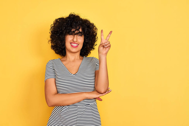 Jeune femme arabe aux cheveux bouclés portant une robe rayée sur fond jaune isolé souriant avec un clin d'oeil heureux au visage de la caméra faisant signe de victoire. Numéro deux
. - Photo, image