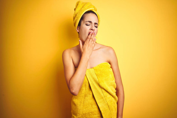 Beautiful woman wearing shower towel on body and head over yellow isolated background bored yawning tired covering mouth with hand. Restless and sleepiness. - Photo, Image