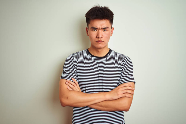 Молодой азиатский китаец в полосатой футболке стоял на изолированном белом фоне скептически и нервно, неодобряя выражение лица со скрещенными руками. Отрицательное лицо
. - Фото, изображение