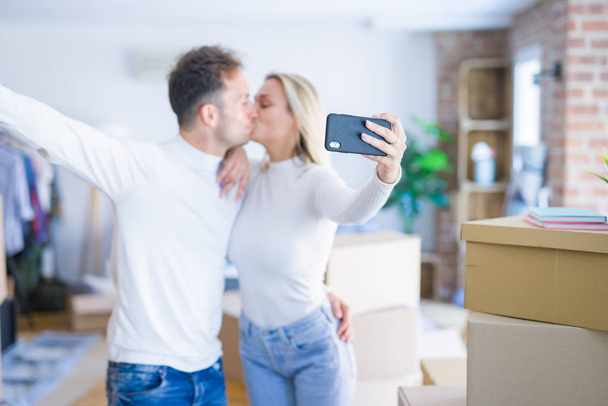 junges schönes Paar steht mit Smartphone für Selfie-Küsse im neuen Zuhause um Kartons herum - Foto, Bild
