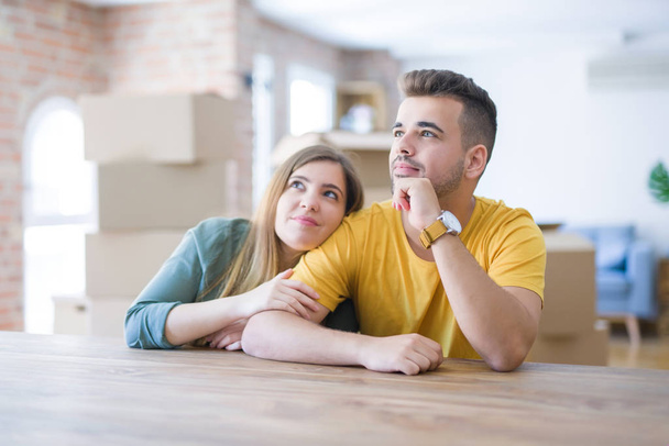 Young couple sitting on the table movinto to new home with carboard boxes behind them with hand on chin thinking about question, pensive expression. Smiling with thoughtful face. Doubt concept. - Photo, Image