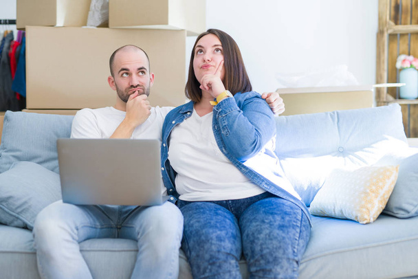 Casal jovem sentado no sofá arround caixas de papelão que se deslocam para uma nova casa usando laptop rosto sério pensando em questão, ideia muito confusa
 - Foto, Imagem