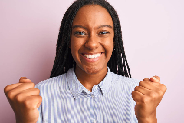 若いアフリカ系アメリカ人女性がピンクの背景の上に立って縞模様のシャツを着て誇りと勝利と成功を祝う叫んで非常に興奮し、感情を応援 - 写真・画像