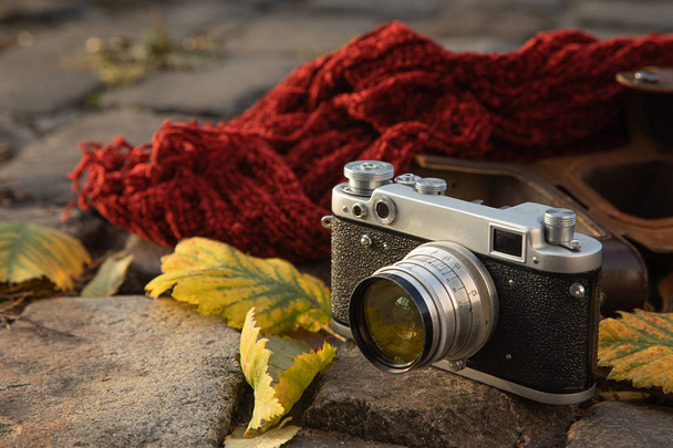 Sonbahar ruh hali natürmort: vintage rangefinder kamera, kahverengi deri kılıf, kırmızı eşarp ve kaldırımda döşeme yaprakları - Fotoğraf, Görsel