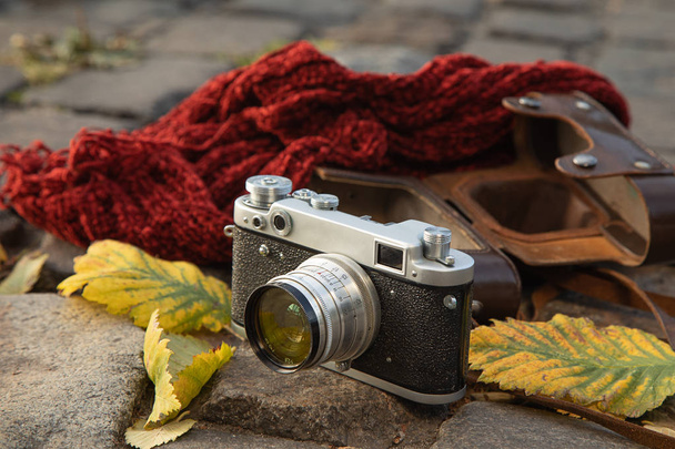 Autumn mood still life: vintage rangefinder camera, brown leather holster, red scarf and leaves laying on pavement - Photo, Image