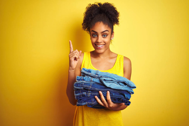 Jonge Afro-Amerikaanse vrouw die spijkerbroeken vasthoudt over geïsoleerde gele achtergrond verrast met een idee of vraag wijzend met een vrolijk gezicht, nummer één - Foto, afbeelding