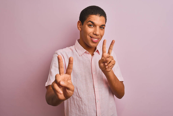 junger gutaussehender arabischer Mann in lässigem Hemd, der vor isoliertem rosa Hintergrund steht und lächelnd in die Kamera schaut und Finger zeigt, die Siegeszeichen machen. Nummer zwei. - Foto, Bild