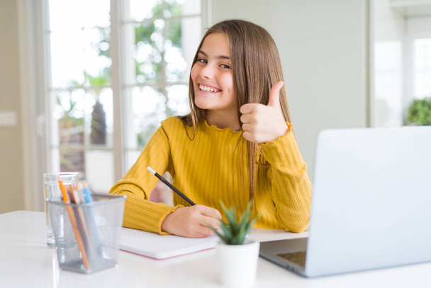 美しい若い女の子は、コンピュータのラップトップを使用して勉強し、大きな笑顔で幸せなノートブックに書きますokサインをします,指で親指アップ,優れたサイン - 写真・画像