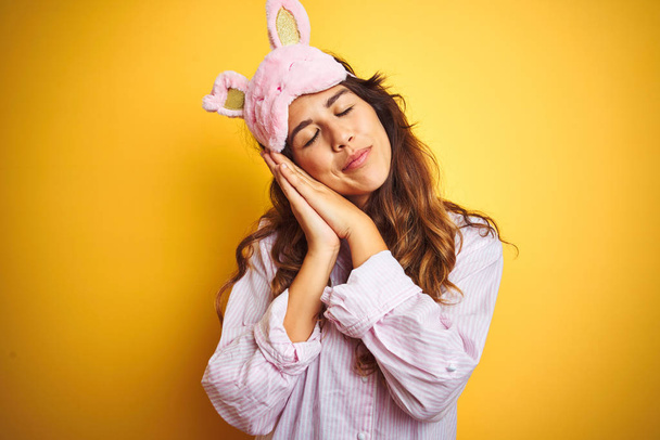 Fiatal nő visel pizsama és alvás maszk felett áll sárga elszigetelt háttér alszik fáradt álmodozás és pózol a kezét, miközben mosolyogva csukott szemmel. - Fotó, kép