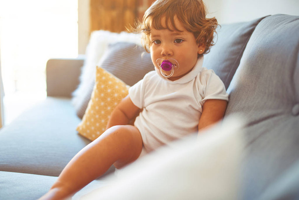 Hermosa niña pequeña con camiseta blanca sentada en el sofá usando chupete
 - Foto, Imagen