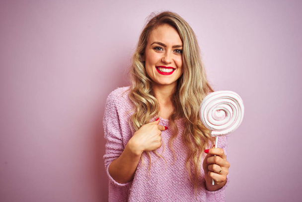 Jovem bela mulher comendo doces doces sobre rosa isolado fundo com surpresa rosto apontando dedo para si mesmo
 - Foto, Imagem