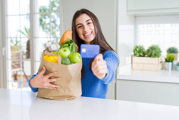 Young woman holding a paper bag full of fresh groceries and showing credit card as payment metod - Foto, afbeelding