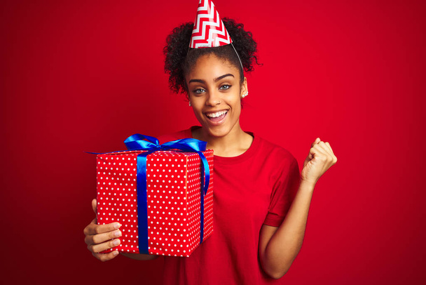 アフリカ系アメリカ人の女性は、孤立した赤い背景の上に誕生日プレゼントを保持面白い帽子を身に着けている誇りと勝利と成功を祝う非常に興奮し、応援感情 - 写真・画像