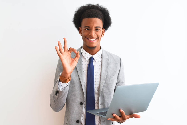 Молодой африканский американский бизнесмен, используя ноутбук стоя на изолированном белом фоне делает хорошо знак с пальцами, отличный символ
 - Фото, изображение