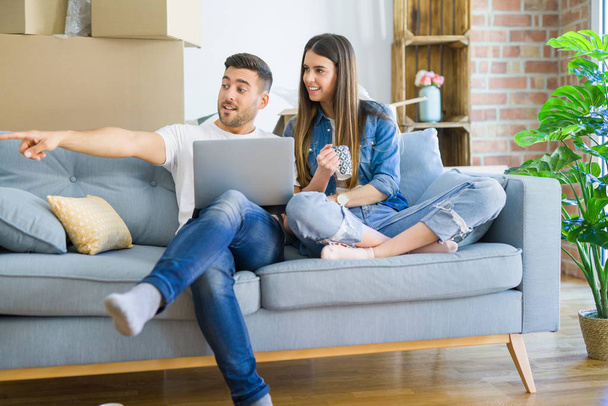 Νεαρό ζευγάρι που κινείται σε ένα νέο σπίτι χαλαρωτικό καθιστός στον καναπέ χρησιμοποιώντας φορητό υπολογιστή, χαμογελαστός χαρούμενος για τη μετακίνηση σε νέο διαμέρισμα - Φωτογραφία, εικόνα