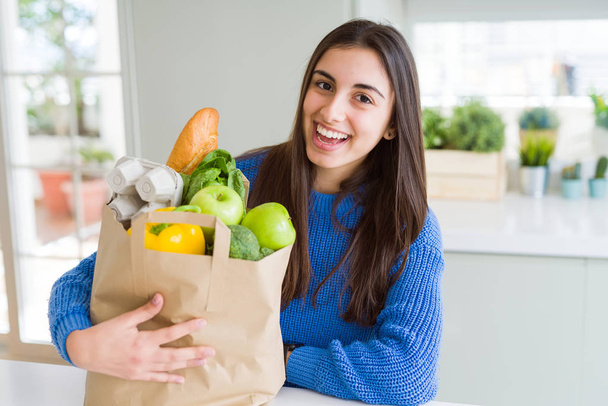 Młoda piękna kobieta uśmiechnięta trzyma papierową torbę pełną artykułów spożywczych w domu - Zdjęcie, obraz