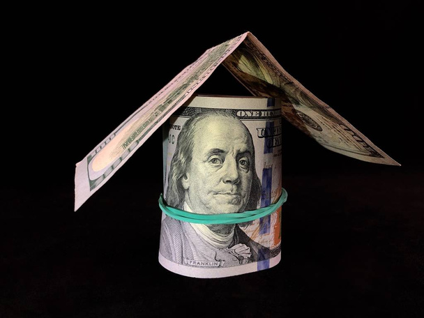 Bankbiljetten van 100 Us dollars zijn verdraaid met een elastische band. Amerikaans geld wordt opgestapeld in de vorm van een huis. Afbeelding van president Franklin op dollars, op een zwarte achtergrond. - Foto, afbeelding